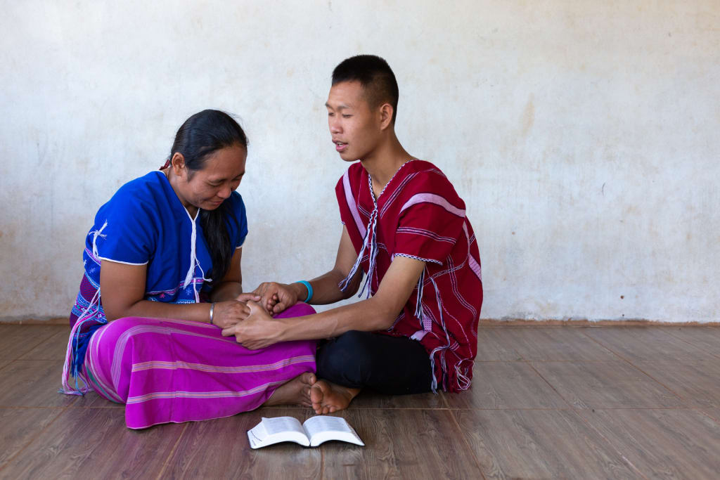 A mother from Thailand holds her teenage sons hands as they pray together seated on the floor with a bible open in front.