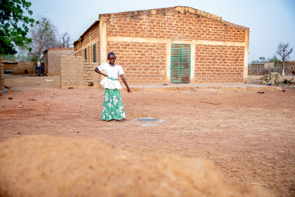 A girl in white top and green skirt stands in front of a building and points at a hole in the ground.