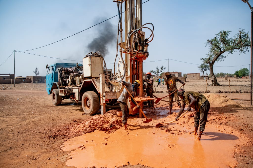 Three men with shovels work beside a machine that is drilling into the earth.