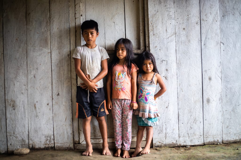A boy and two girls stand in front of a white wall and look solemn.