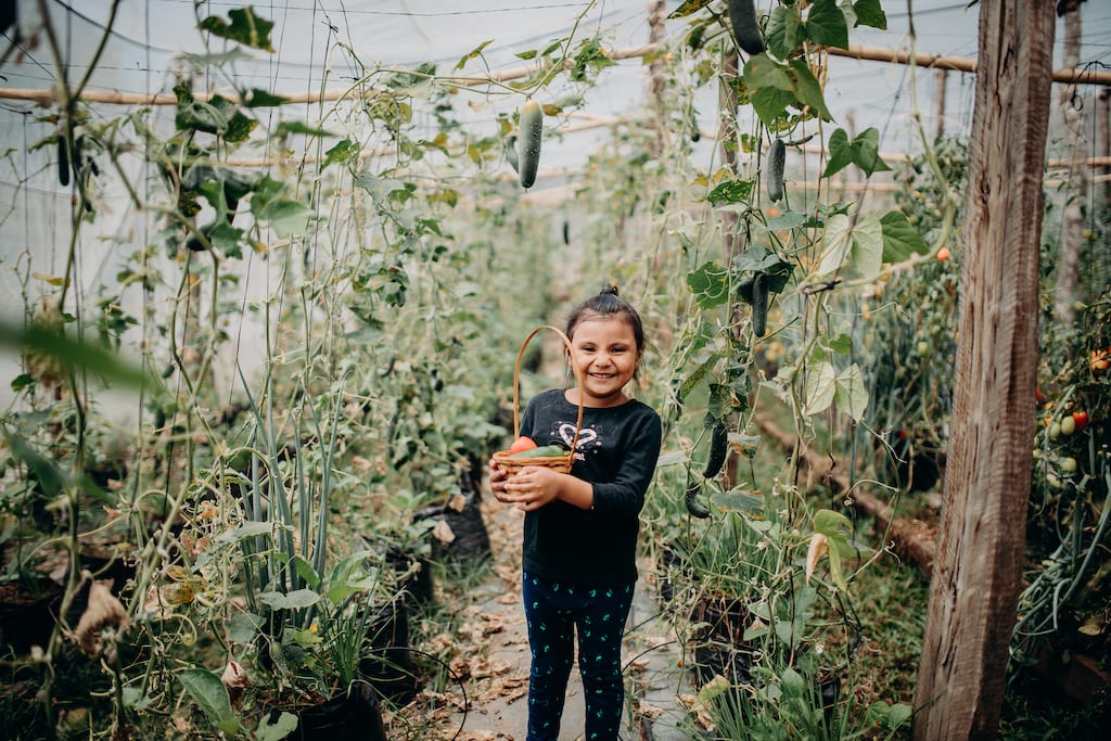 Daniela, in a black shirt, is smiling and standing in a vegetable garden holding a basket of vegetables she has collected.