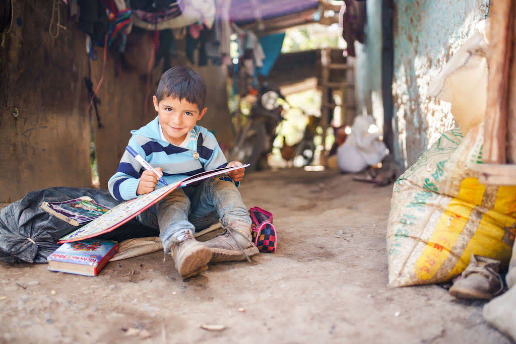 Boy sits on the ground writing in a book and smiling.