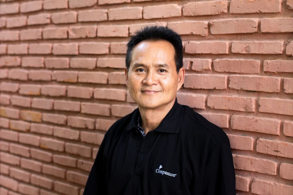 A man in a black shirt smiles at the camera in front of a brick wall