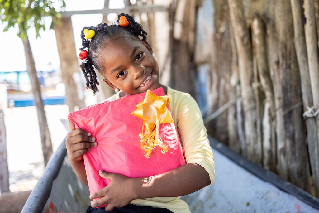 In Colombia, Zahen is wearing a yellow shirt. She is sitting in a boat on the seashore. She is holding her Christmas gift from the center, which is wrapped in pink paper with a yellow bow.