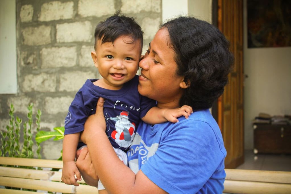 Jeinny is wearing a blue shirt. She is holding her son, Argya, wearing a dark blue shirt, outside their home.