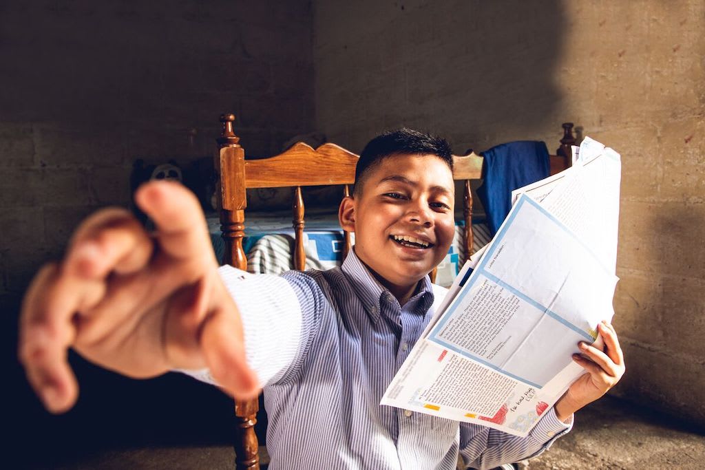 Josué is wearing a blue and white striped shirt. He is sitting in a wooden chair inside his home and is holding letters from his sponsor.