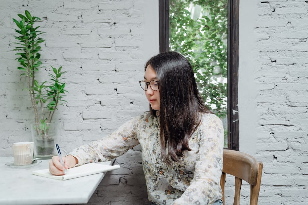 Girl wearing floral shirt and glasses writes with a pen at her desk against a white brick wall