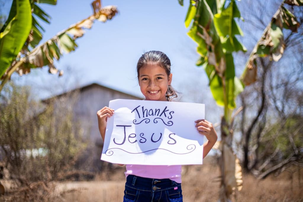 Little girl wearing a purple shirt stands in front of two trees holding a sign that says, "Thanks Jesus"