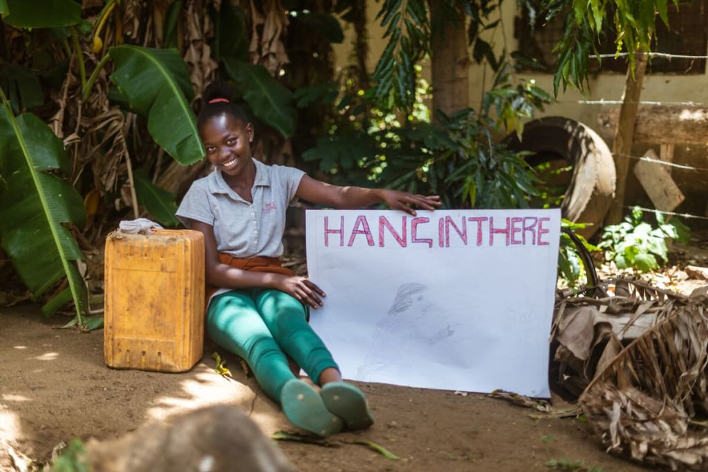 Girl sits under a tree holding a sign that says, "Hang in there"