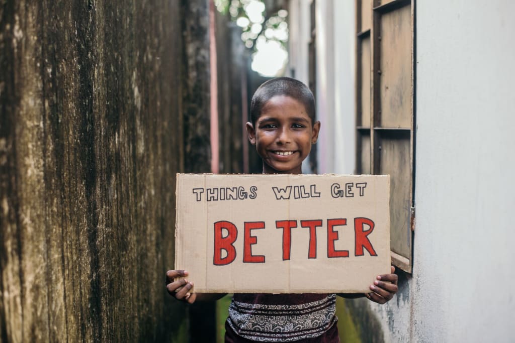 A little boy stands in an alley way holding a sign that says "Things will get better."