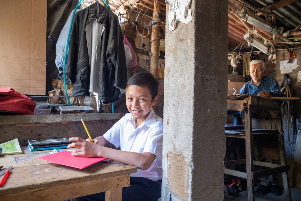 Eleazar sits at a table in his home doing schoolwork. His dad sits at a table in the background.