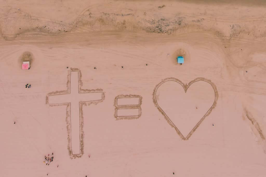 A "cross equals heart" symbol written in sand on a beach.