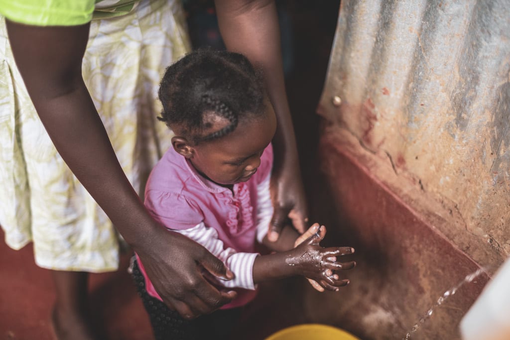 A young toddler girl washes her hands with the help of her mother.