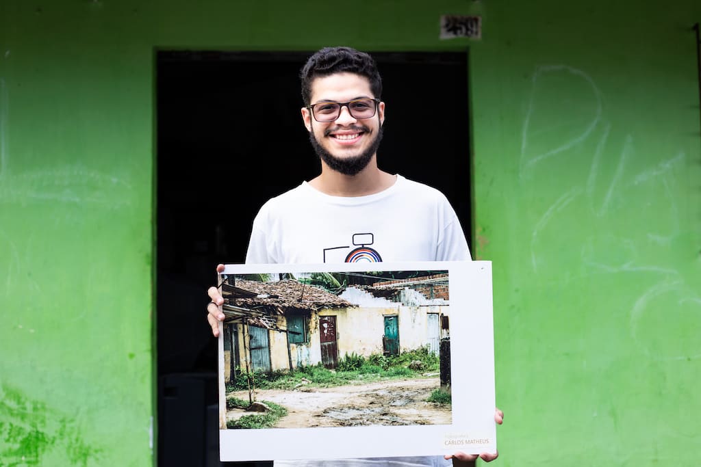 Matheus stands in front of a green building, holding a photograph he took of a home.