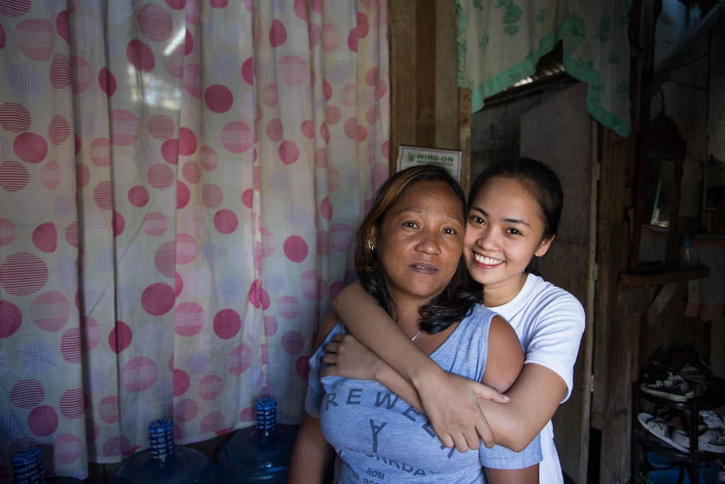 Maricris stands behind her mother with her arms around her. Maricris is wearing a white t-shirt and smiling, her mother is wearing a light purple t-shirt and has a neutral look on her face.