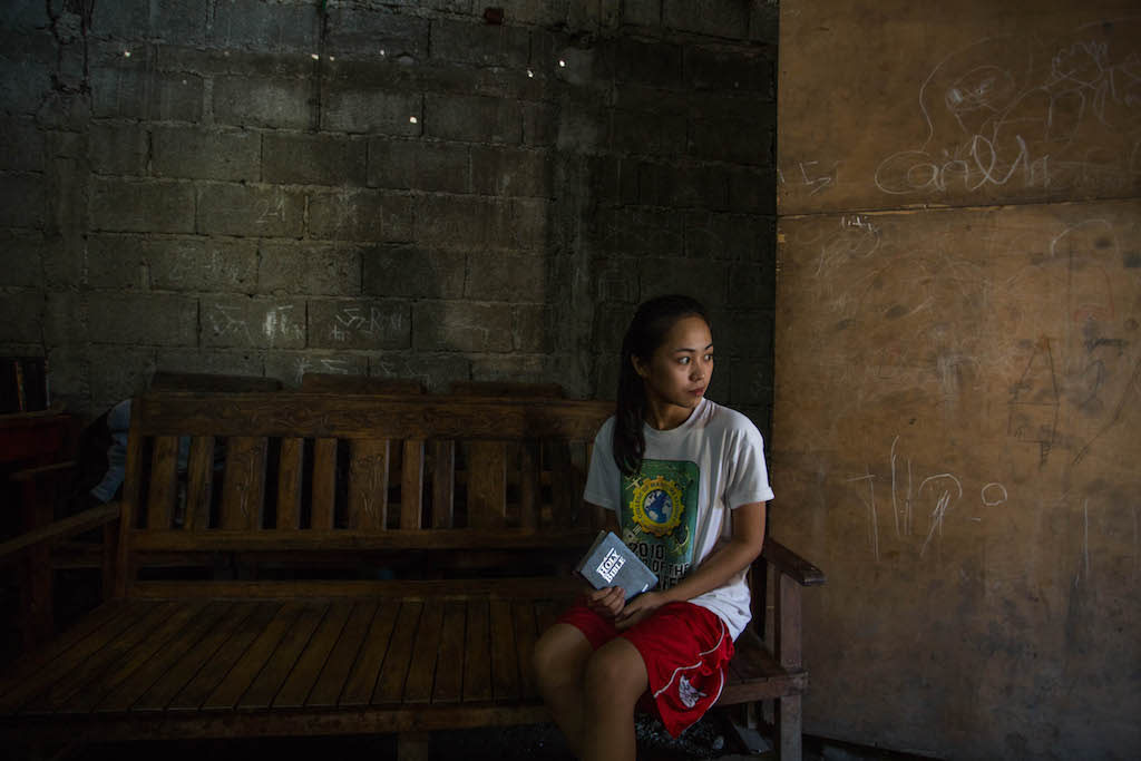 Maricris sits on a wooden bench, looking to the side. She wears a white tshirt and red shorts, and she is holding a Bible. Her long black hair is tied in a ponytail.
