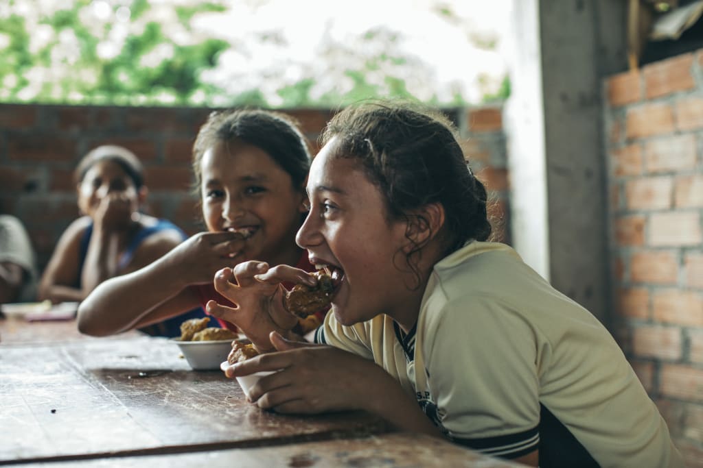 A girl sits at a table eating some chicken out of a bowl. She and her friends are smiling.