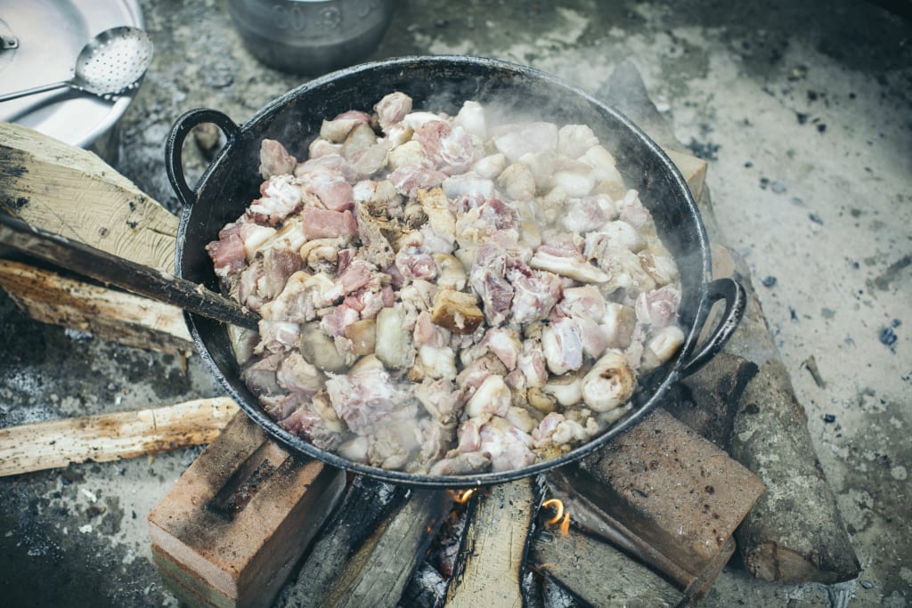 A large black pan cooks meat over an open fire.