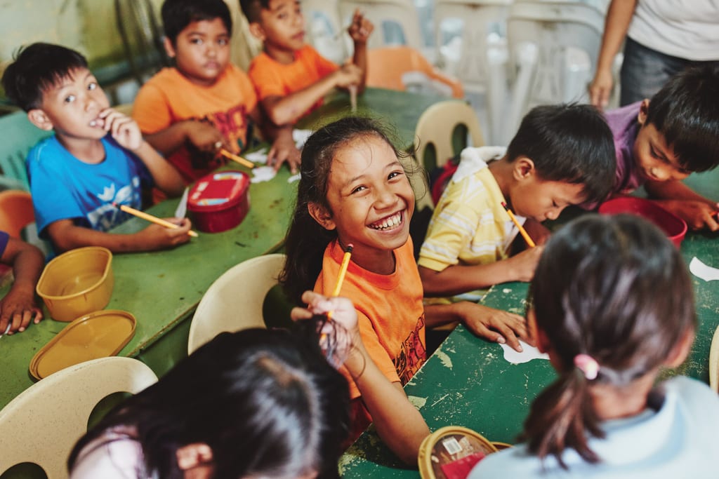 A Filipino girl looks up from sitting at a table, smiling and wearing an orange teenager. Other engaged children surround her.
