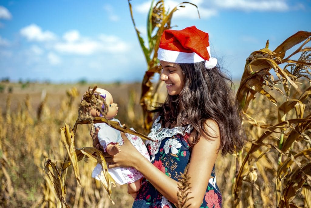 A little girl holds a new doll and wears a Santa hat. She is standing in a corn field.