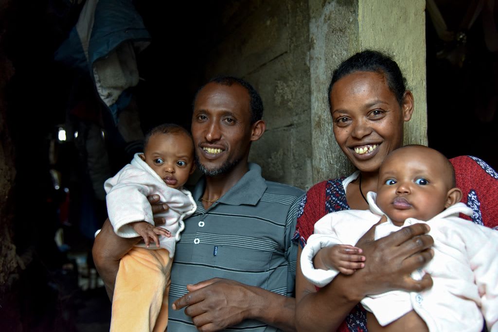 A mom and dad hold their two babies. They are smiling in the doorway of their home.
