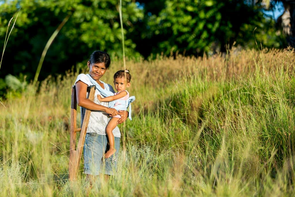 A mother, wearing a baby carrier in her shoulders, walks in a tall grass countryside with her wood crutch, holding a baby, in her arms, posing outside with her child and green trees in the background.