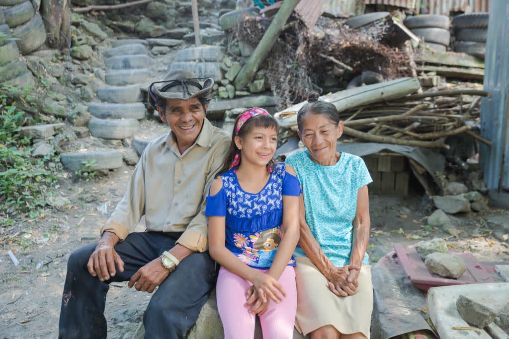A girl, wearing a blue shirt, pink headband in her hair, and pink pants, sits outside on a large rock with her grandparents near their home.