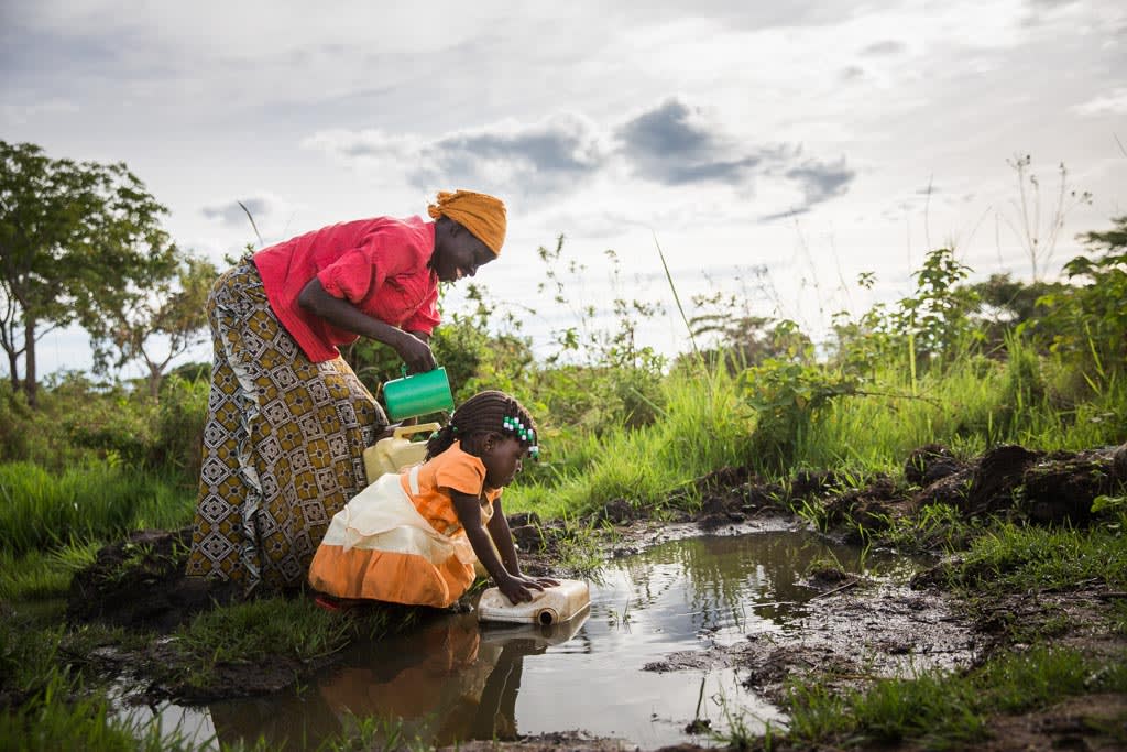 Mother and child fetch water behind their home. Betty, a girl wearing an orange and white dress, bends over to fetch water with a plastic container beside her mother as they are getting water in plastic buckets and containers. Evelyn, who is wearing a yellow scarf, turban, head wrap on her head, a red shirt and long pattern skirt, together at a large mud puddle of collected rain water on the ground, with the mother using a green cup in her hands to retrieve the water to pour into the containers. This appears to be an unsanitary water source, potentially unhealthy, unsafe, and not clean drinking water in a large puddle on the ground. They are surrounded by green trees and grass.