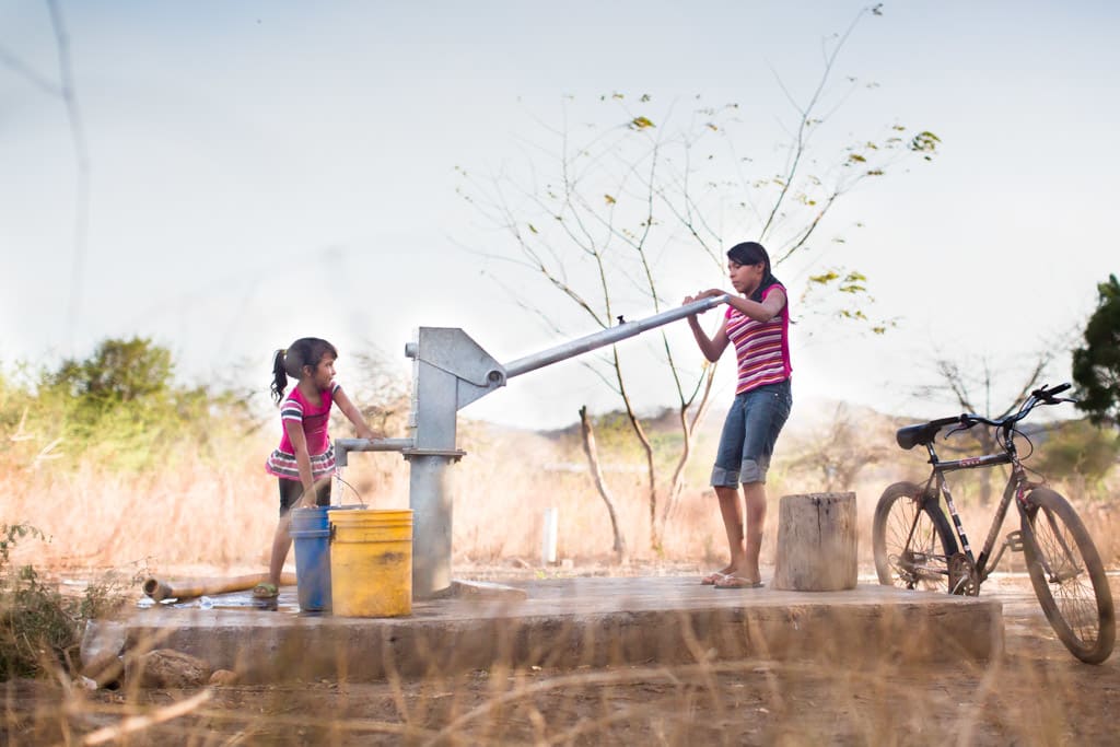 Two children working together to get water from a well. A bicycle is to the right of them and they are in a field.