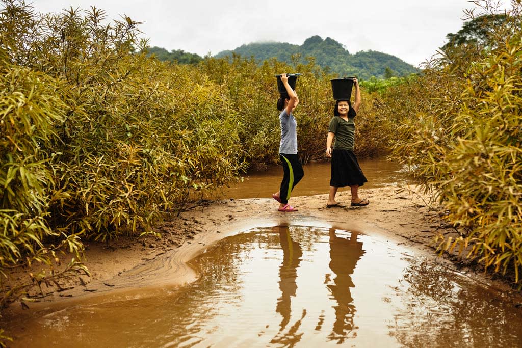Two girls are pictured carrying water buckets on their heads by a river. They are smiling and surrounded in trees.
