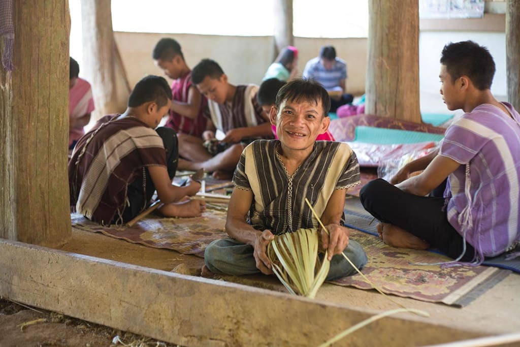 A man in Thailand sits on the ground weaving a basket alongside students.