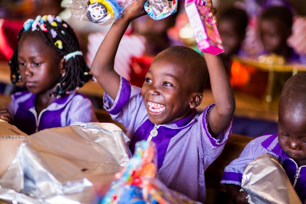 I little boy in Burkina Faso sits in class, opening his christmas presents. He looks very excited, smiling as he holds his new toys above his head.