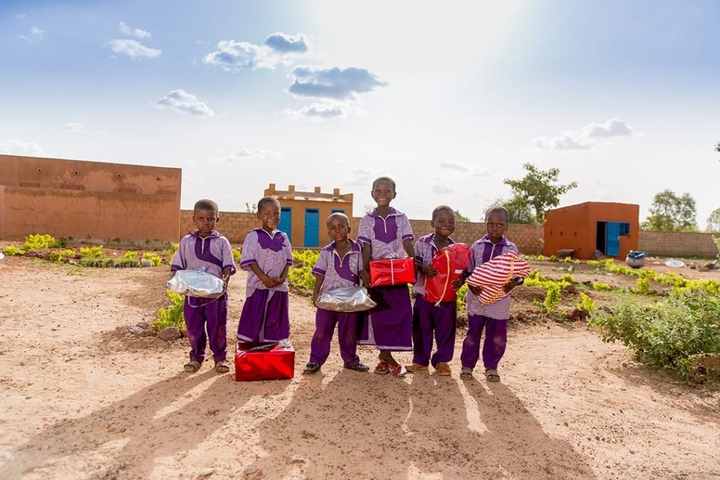 Six children from Burkina Faso stand in front of their Compassion centre. It has brown walls and vibrant, blue doors. They were ornate, purple school uniforms and hold Christmas presents