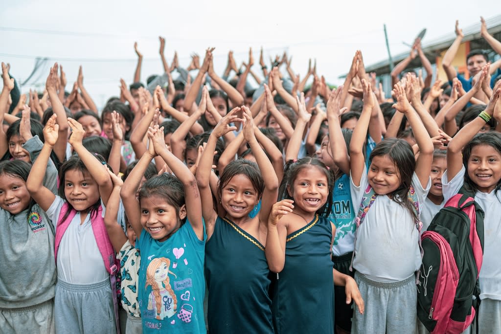 A group of kids in Ecuador are all smiling and clapping with their hands in the air.