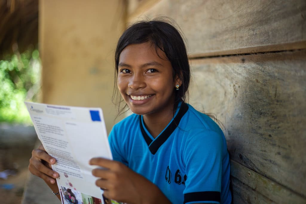 A Colombian girl in a blue t-shirt smiles as she reads a letter from her sponsor.