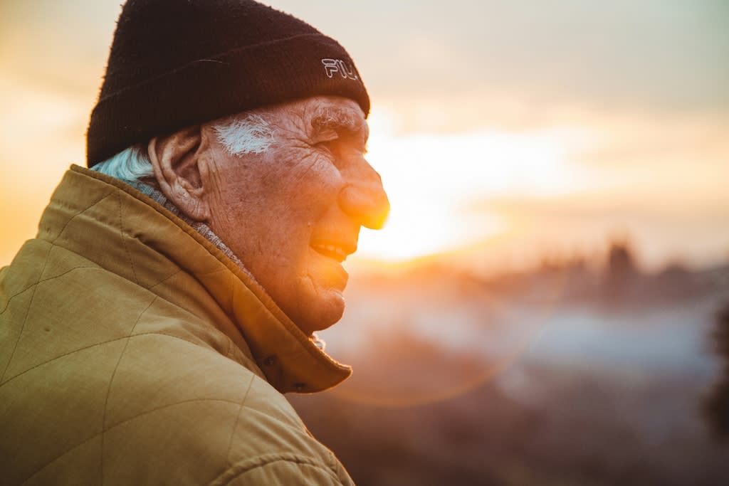 A close-up of an elderly man with a toque and a beige jacket looks out into the setting sun.