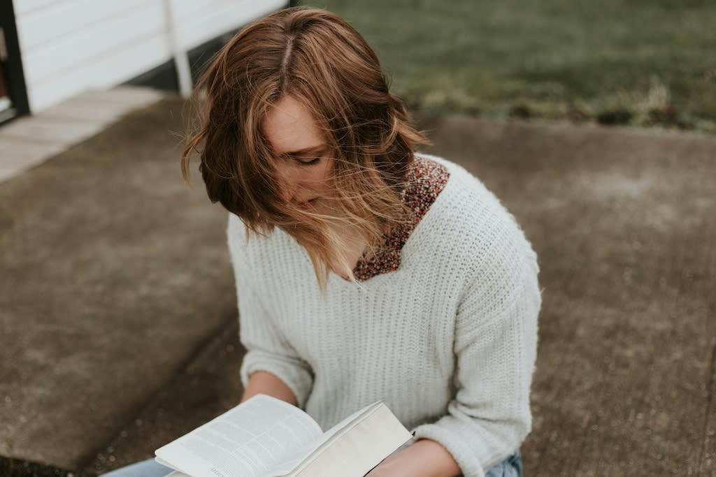 A woman with red hair is wearing a white knitted sweater and reading a bible in her lap.