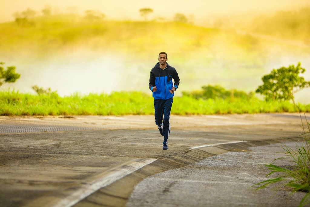 A man is running down a road with a blue jacket and hills in the background.