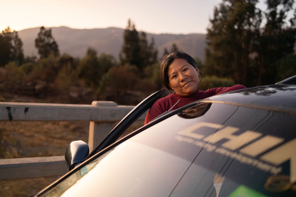 Rosemary stands on the other side of a taxi and smiles. The sun is hitting the windshield.