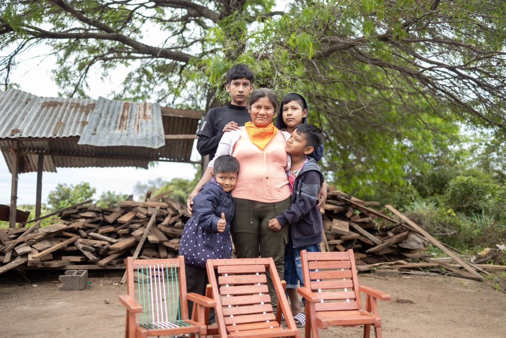 A woman is posing for a picture with her children. In front of them are three of the chairs Elena made. There is a wood pile in the background.
