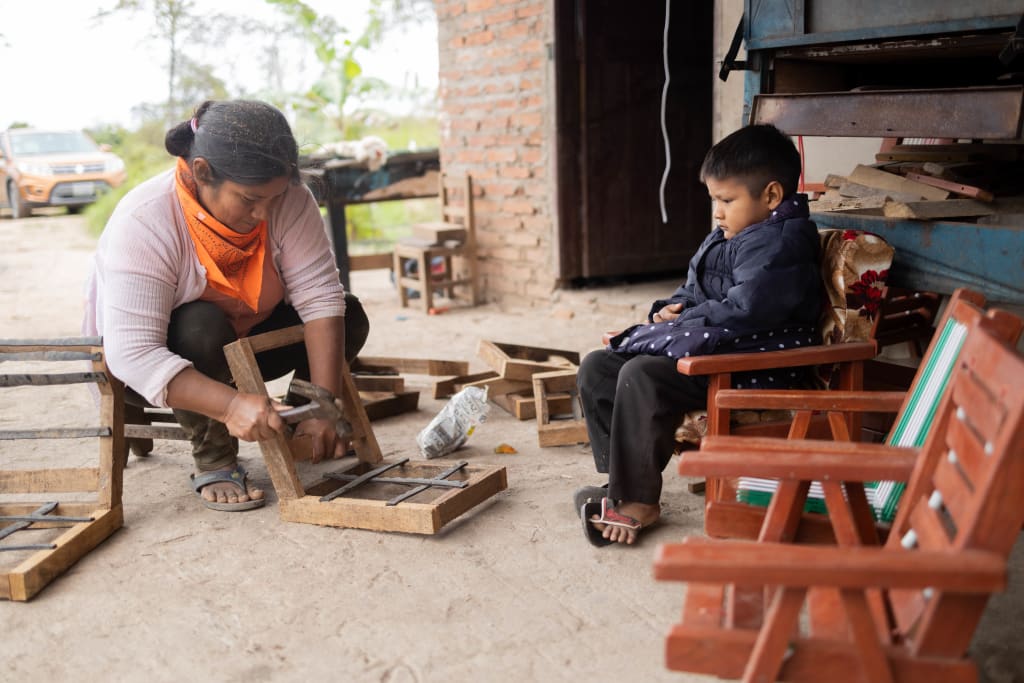 A woman bends over while working with some tools. Her son sits in a small chair watching her.