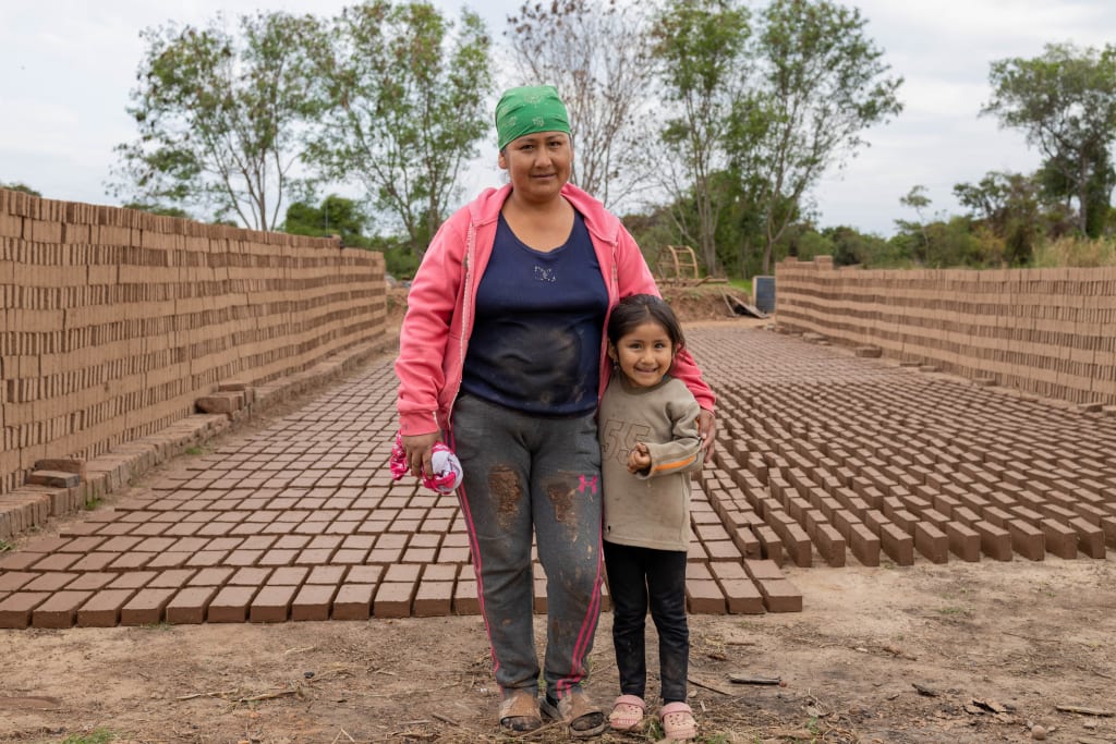 A woman is standing with her young daughter. There are rows of bricks that Janeth made on the ground behind them.