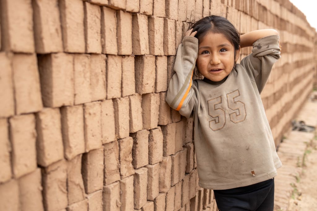 Kimey plays while her mother, Janeth, works making bricks. The girl is leaning against a brick wall.