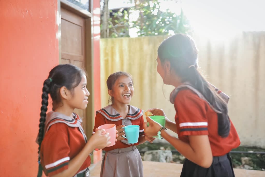 3 girls holding cups talk and laugh with each other outside of a school. They are wearing orange uniforms.