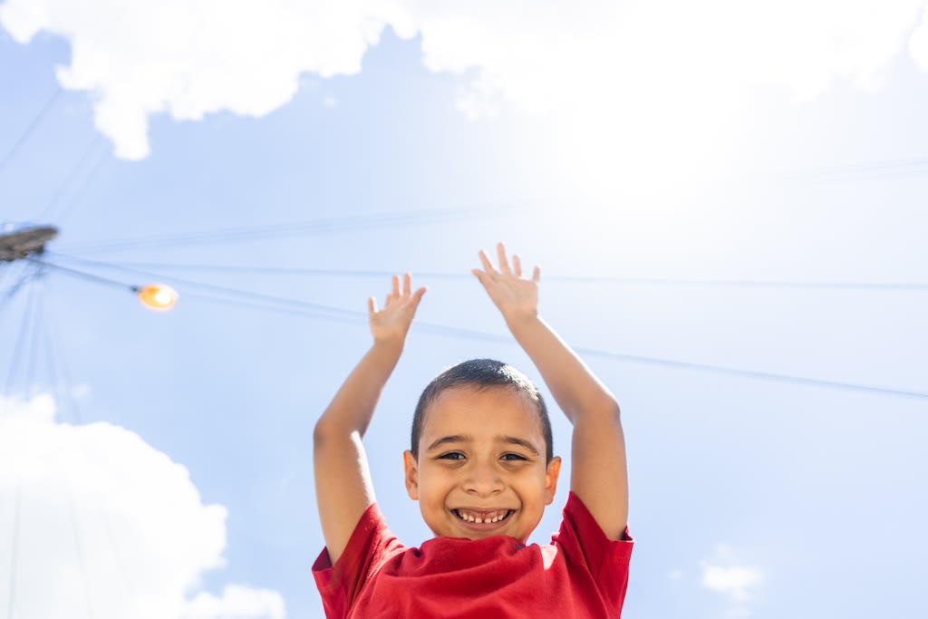 A little boy smiles and holds his hands high in the air. You can see the blue sky and clouds.