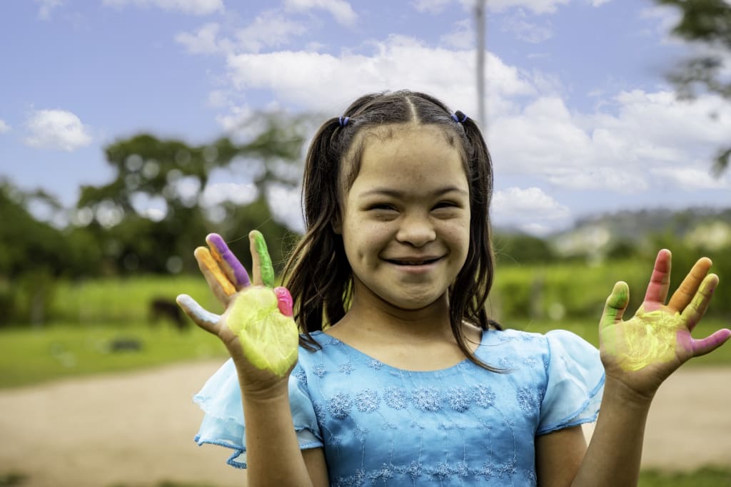 Girl wears a blue dress and smiles holding up her hands that are covered in paint.