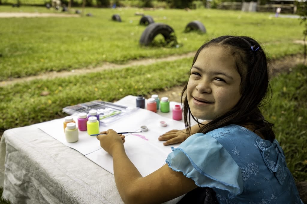 Girl wears a blue dress and paints on paper in a field. She is turning around to smile at the camera.