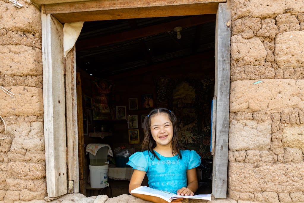Girl with a blue dress smiles at the camera reading a book in the doorway in her home.