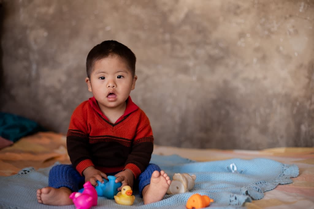 A baby boy sits on a blue blanket with some colourful toy ducks. He is wearing a red sweater.