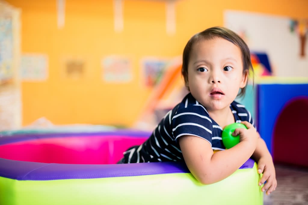 A baby girl wearing a striped blue t-shirt. She is in a play pool holding a toy ball.